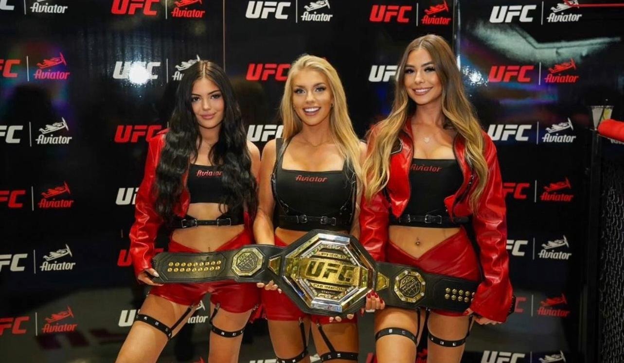 UFC ring girls posing with championship belt in front of UFC and Aviator-branded backdrop