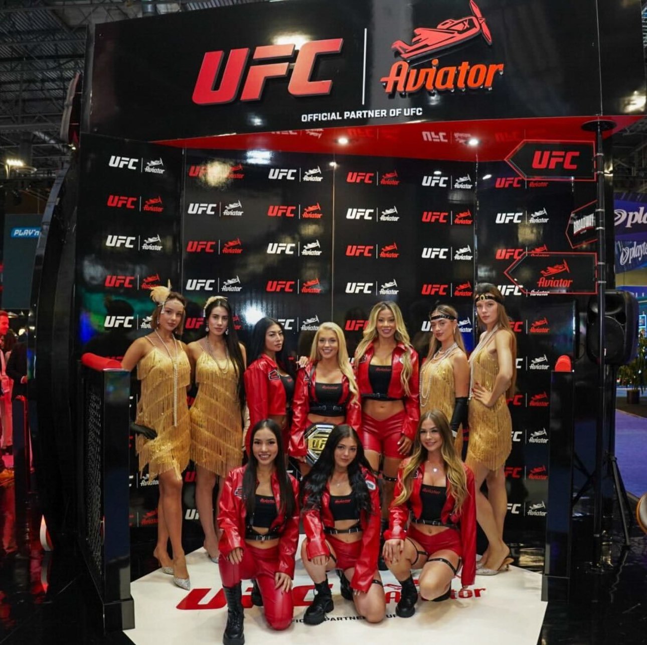 Group of UFC ring girls in red and gold outfits posing at a UFC and Aviator-themed booth