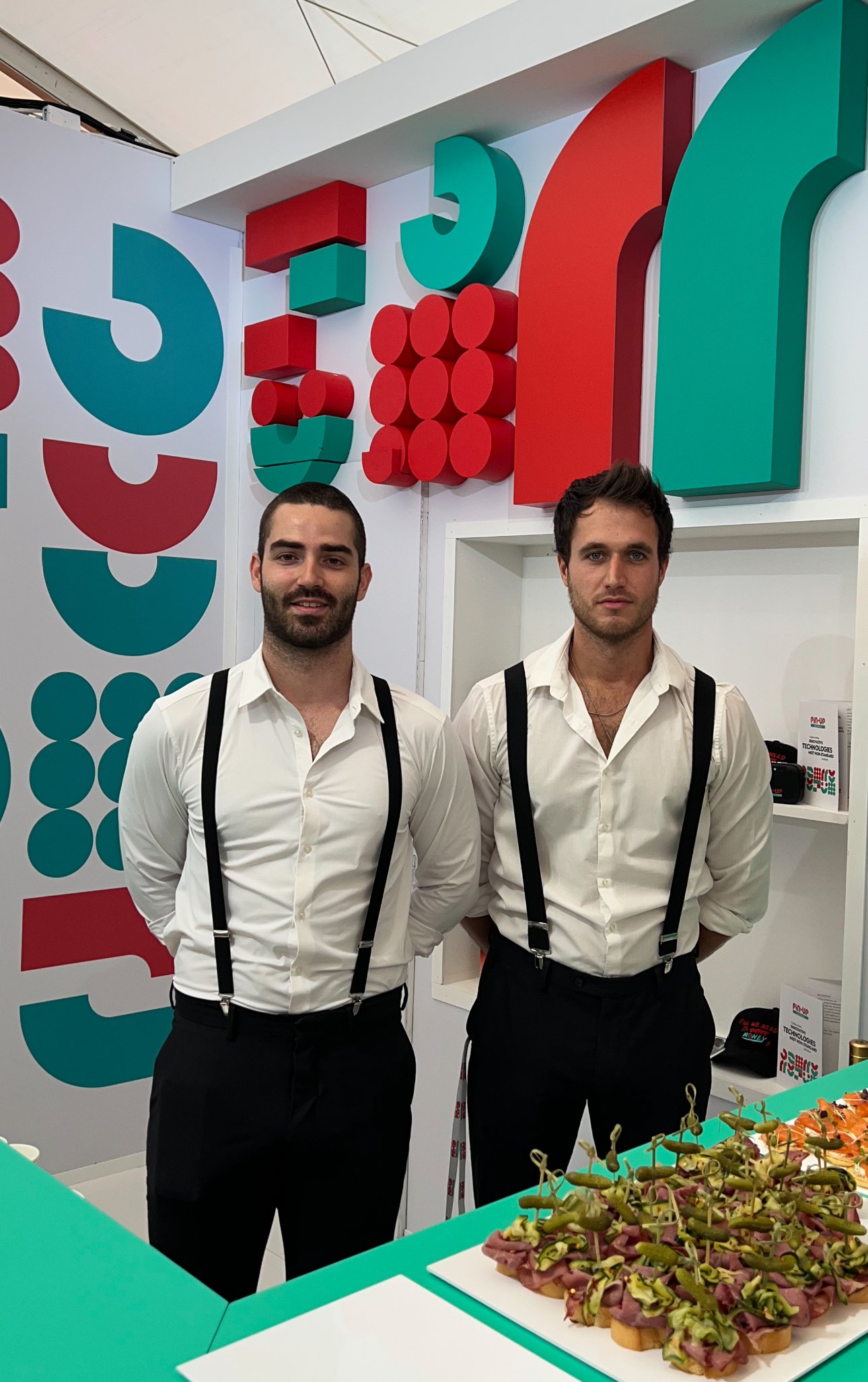 Two men in white shirts with suspenders posing at a vibrant event booth with decorative background