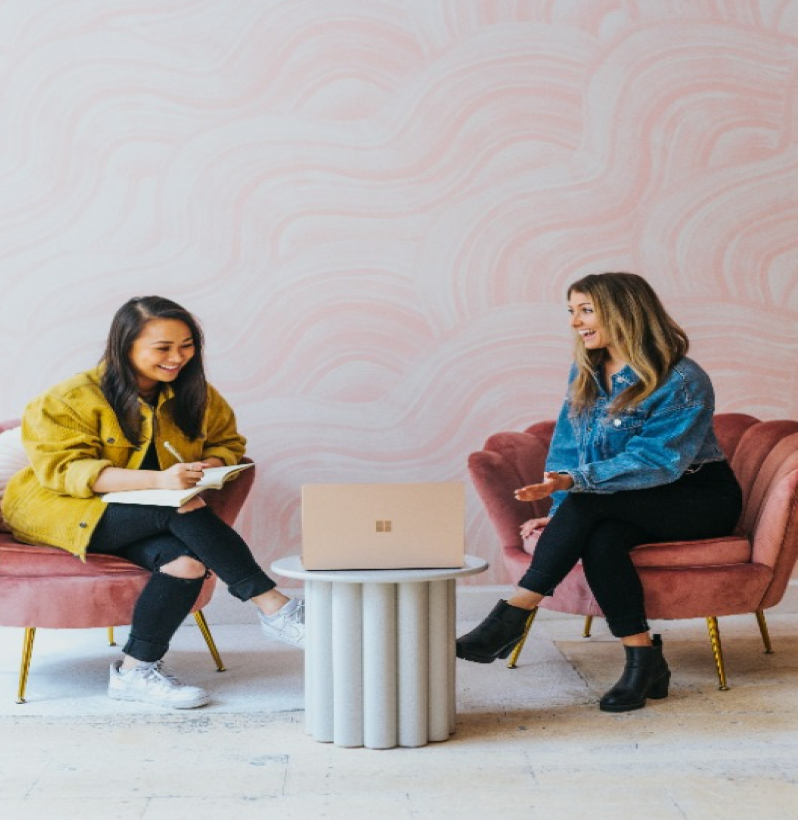 Two women having a friendly discussion in a cozy office lounge with pink chairs